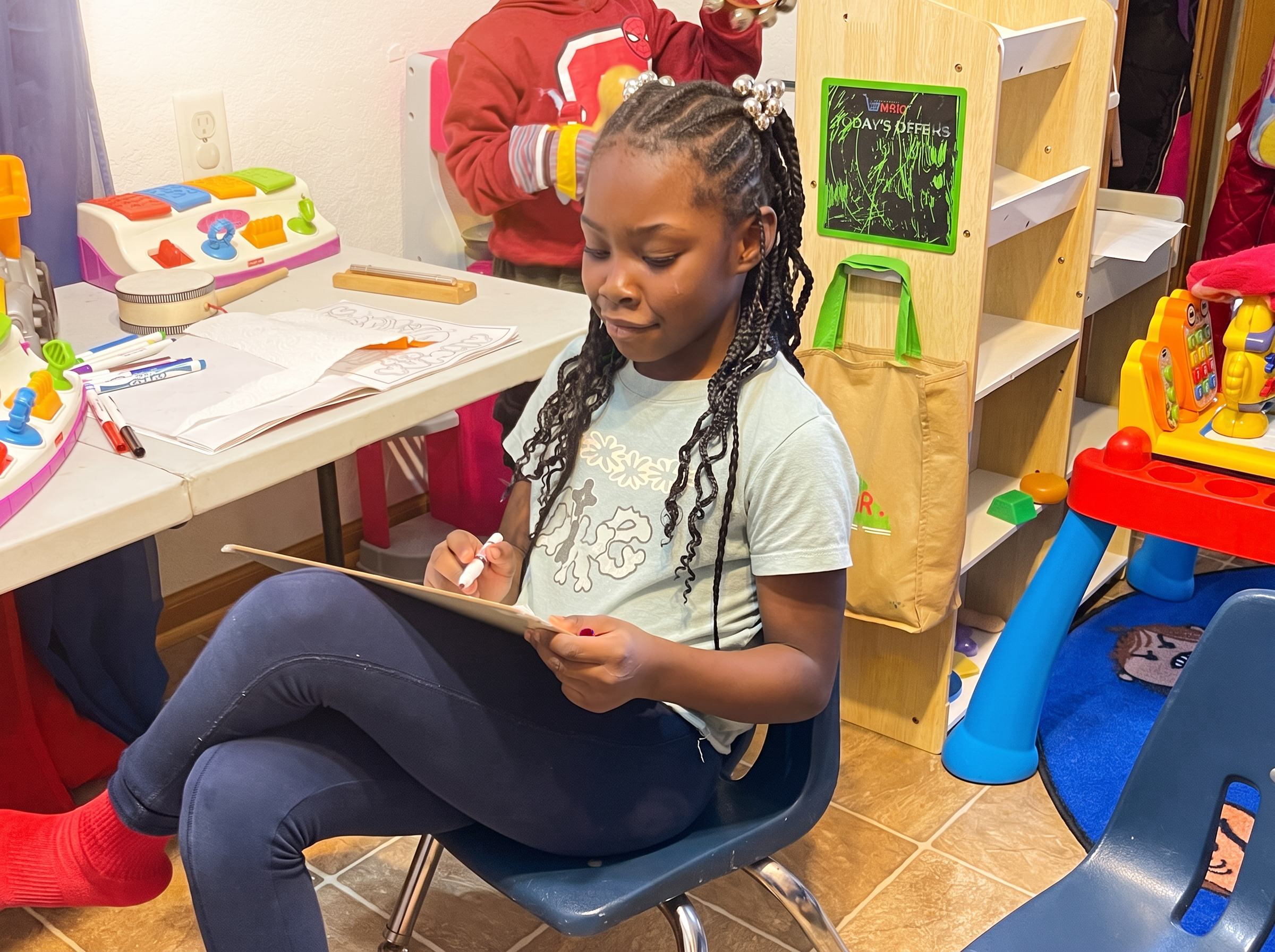Child coloring at a classroom table surrounded by toys and learning materials